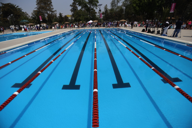 Großes Schwimmbad mit Bahnentrennern, umgeben von Menschen mit Taschen und Gegenständen, mit Zelten, Schildern, Stangen, einem Schild und einer bewölkten Himmel im Hintergrund.