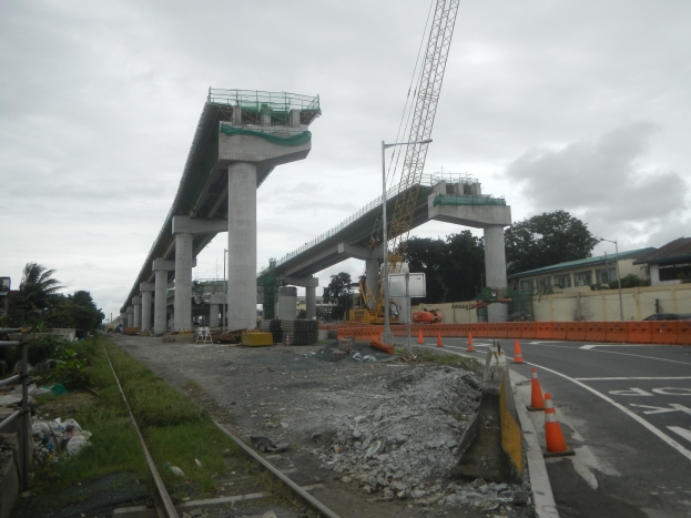 Baustelle mit einer Brücke im Hintergrund, Straße mit Verkehrskegeln markiert, Schienenstraße auf der linken Seite, verstreute Steine und Gras, Bäume und Gebäude säumen die Straße und ein bewölkter Himmel.