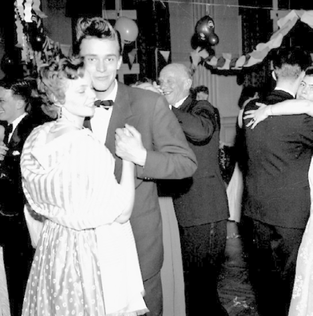 Ein Schwarz-Weiß-Foto von einem Mann und einer Frau, die auf einer Party tanzen, umgeben von lachenden Zuschauern und mit Ballons und Dekorationen im Hintergrund.