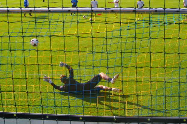 Ein Fußballspieler taucht ab, um einen Ball vor einem Tor zu halten, mit einem Netz im Vordergrund und einigen Menschen im Hintergrund, während mehrere Fußballschuhe auf dem Boden verstreut sind.