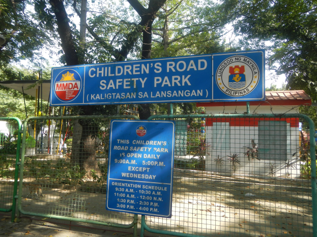 Metallzaun um einen Kinderverkehrssicherheitspark mit zwei blauen Informationsschildern, einem kleinen Haus, Pflanzen, Bäumen und einem bewölkten Himmel.