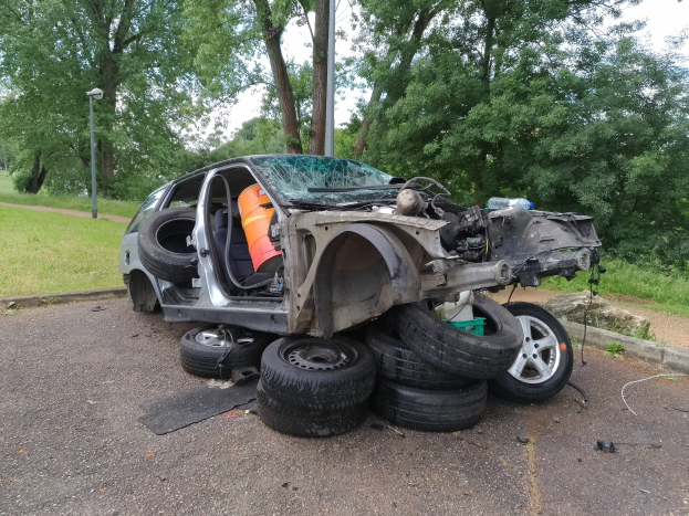Ein Auto liegt auf der Seite am Stra├čenrand, umgeben von Gras, Pflanzen, B├Ąumen, einem Laternenpfahl und einem bew├Âlktem Himmel, mit verstreuter Tr├Ąmmern, die auf einen Unfall hindeuten.
