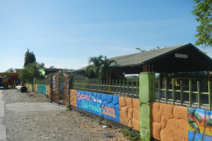 Schulgebäude mit einem bunten Wandgemälde an seiner Seite, umgeben von einem Zaun, neben einem Schuppen und Bäumen, mit parkenden Fahrzeugen, Strommasten und einem bewölkten Himmel.