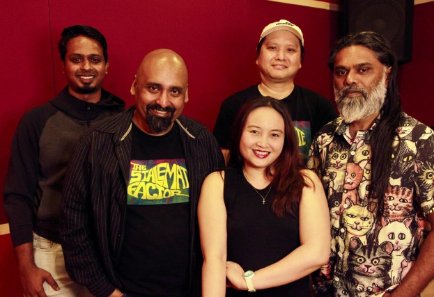 Eine Gruppe von Menschen, die in einem Aufnahmestudio posen, lächeln und leger gekleidet sind, mit einem Lautsprecher im Hintergrund.