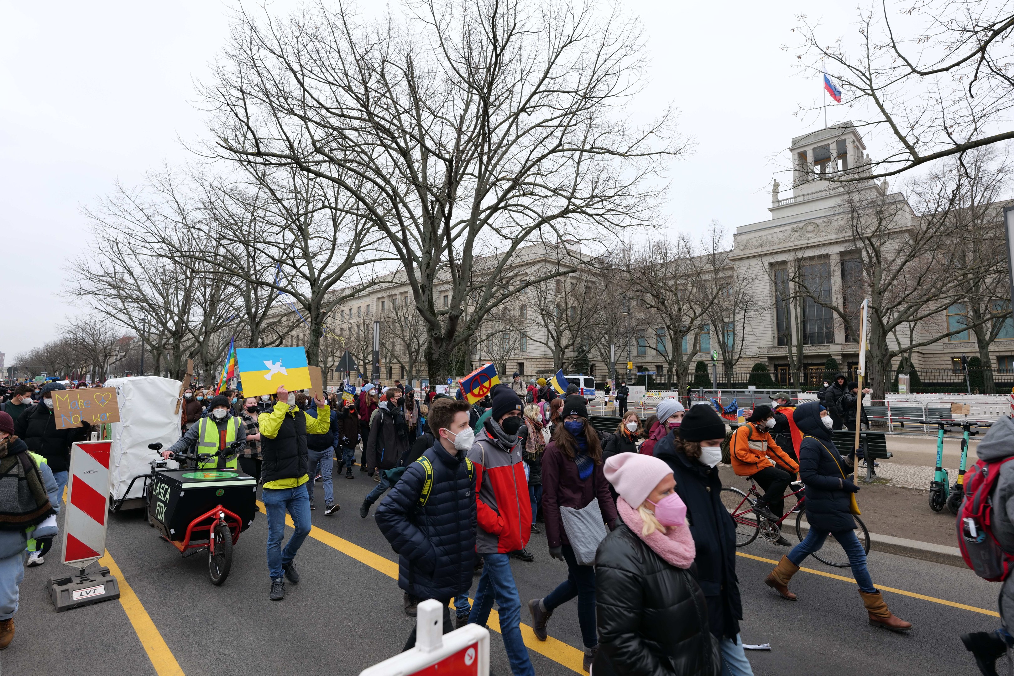 Eine große Protestmarsch mit Menschen, die eine Straße entlanggehen, einige halten Schilder und andere fahren Fahrräder, mit Bäumen und einem klaren blauen Himmel im Hintergrund.
