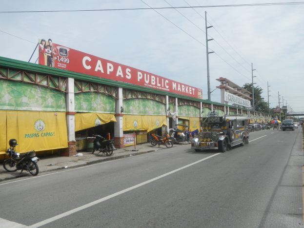 Eine belebte Straße einer Stadt mit Fahrzeugen, einem Gehweg, Strommästen, Gebäuden, Bäumen und einem bewölktem Himmel, mit einem Gebäude mit der Aufschrift "Capas Public Market" im Vordergrund.