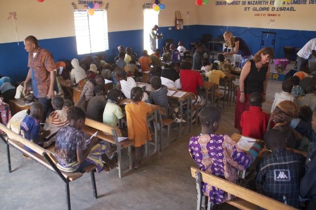 Kinder auf Bänken in der Nähe von Erwachsenen sitzend, während Ballons und Text oben über Möbel im Hintergrund wie Stühle, eine Tür, Fenster und ein Tisch erscheinen.