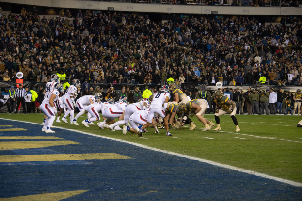 Ein Fußballspiel in vollem Gange mit Spielern in Sportkleidung und Helmen auf dem Rasen, aus der Perspektive eines Stadionplatzes mit Zuschauern, Geländern und Hintergrundwerbung.