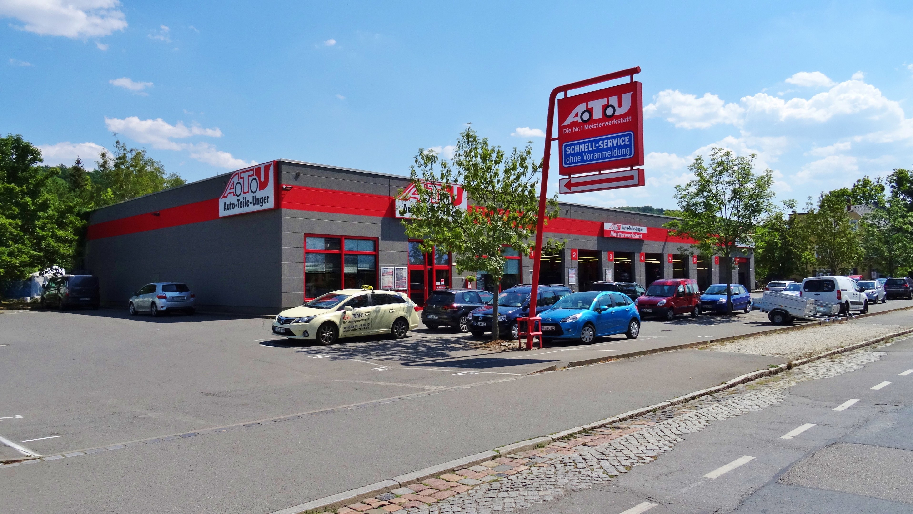 Ein Autohaus mit mehreren geparkten Autos vorne, umgeben von Gebäuden, Bäumen, Strommasten, Schildern und einer Straße, unter einem bewölkten Himmel.