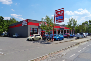 Ein Autohaus mit mehreren geparkten Autos vorne, umgeben von Gebäuden, Bäumen, Strommasten, Schildern und einer Straße, unter einem bewölkten Himmel.