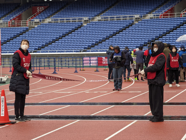 Gruppe von Menschen am Start einer Rennstrecke mit einer Schnur in den Händen, mit einem Verkehrskegel links und Event-Aufbau im Hintergrund.