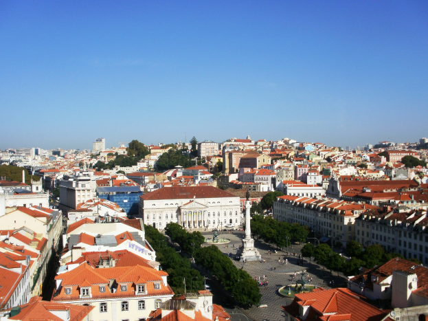 Blick auf Lissabon von einem Hügel aus, der Gebäude, Bäume, eine Statue auf einem Sockel, Menschen auf einer Straße und den Himmel im Hintergrund zeigt.