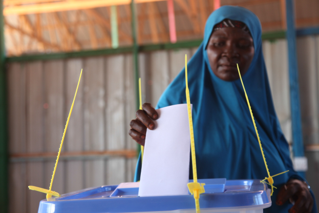 Frau in blauem Kopftuch hält ein Blatt Papier in der Hand und steht neben einer Kiste mit herausragenden Stöcken an einer Wahllokalwand mit einer Hütte im Hintergrund.