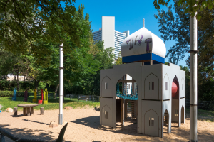 Ein Spielplatz mit einer Spielstruktur in der Mitte, umgeben von Bäumen, Pfählen, Bänken und anderen Gegenständen, mit Gebäuden und einem klaren blauen Himmel im Hintergrund.