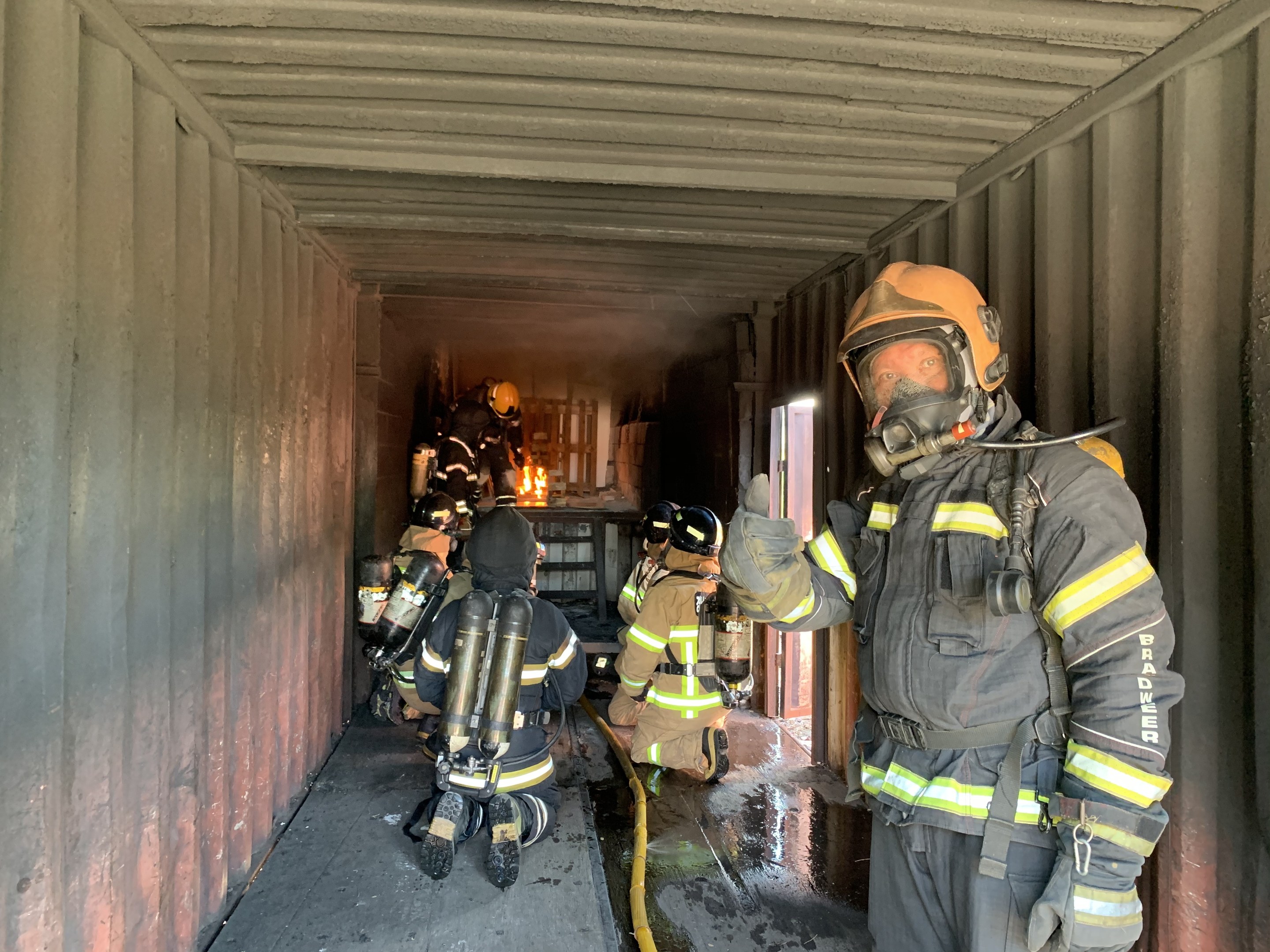Feuerwehrleute in Schutzausrüstung innerhalb eines Containers während eines Feuerlöschvorgangs, einer hält ein Rohr mit Feuerlöscher und Equipment im Hintergrund.