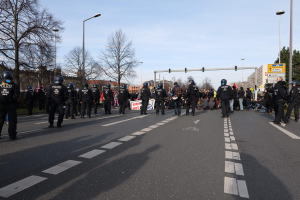 Eine Gruppe von Polizisten in schwarzen Uniformen und Helmen, die an der Seite einer Straße mit Laternen, Ampeln, Bäumen, Gebäuden und einem klaren blauen Himmel im Hintergrund stehen.