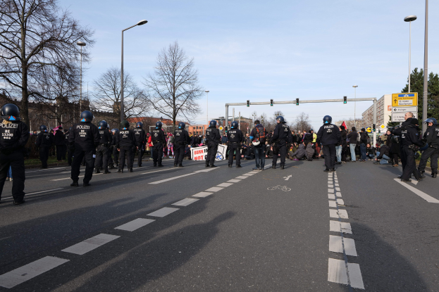 Eine Gruppe von Polizisten in schwarzen Uniformen und Helmen, die an der Seite einer Straße mit Laternen, Ampeln, Bäumen, Gebäuden und einem klaren blauen Himmel im Hintergrund stehen.