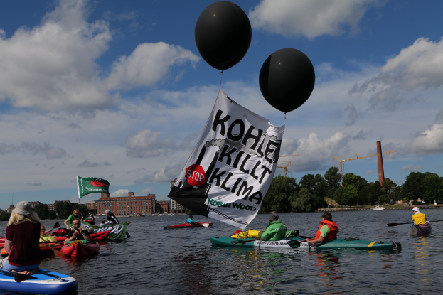 Gruppe von Menschen in Kajaks auf dem Wasser mit einem Banner, das "Kohle Kill Klima" und Bäume, Gebäude und Kräne im Hintergrund unter einem klaren blauen Himmel zeigt.