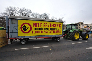 Ein Lastwagen mit einer Aufschrift auf der Rückseite fährt eine Straße entlang, umgeben von Menschen, Bäumen, Gebäuden und einem klaren blauen Himmel, wahrscheinlich Teil einer Demonstration in Deutschland.