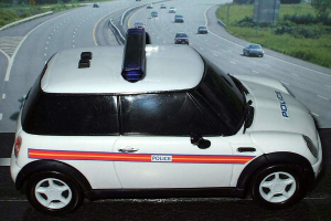 Ein weißer Polizeiwagen mit "Polizei" darauf, der auf einer Straße außerhalb der Stadt fährt, umgeben von Gras, Bäumen und anderen fahrenden Fahrzeugen.