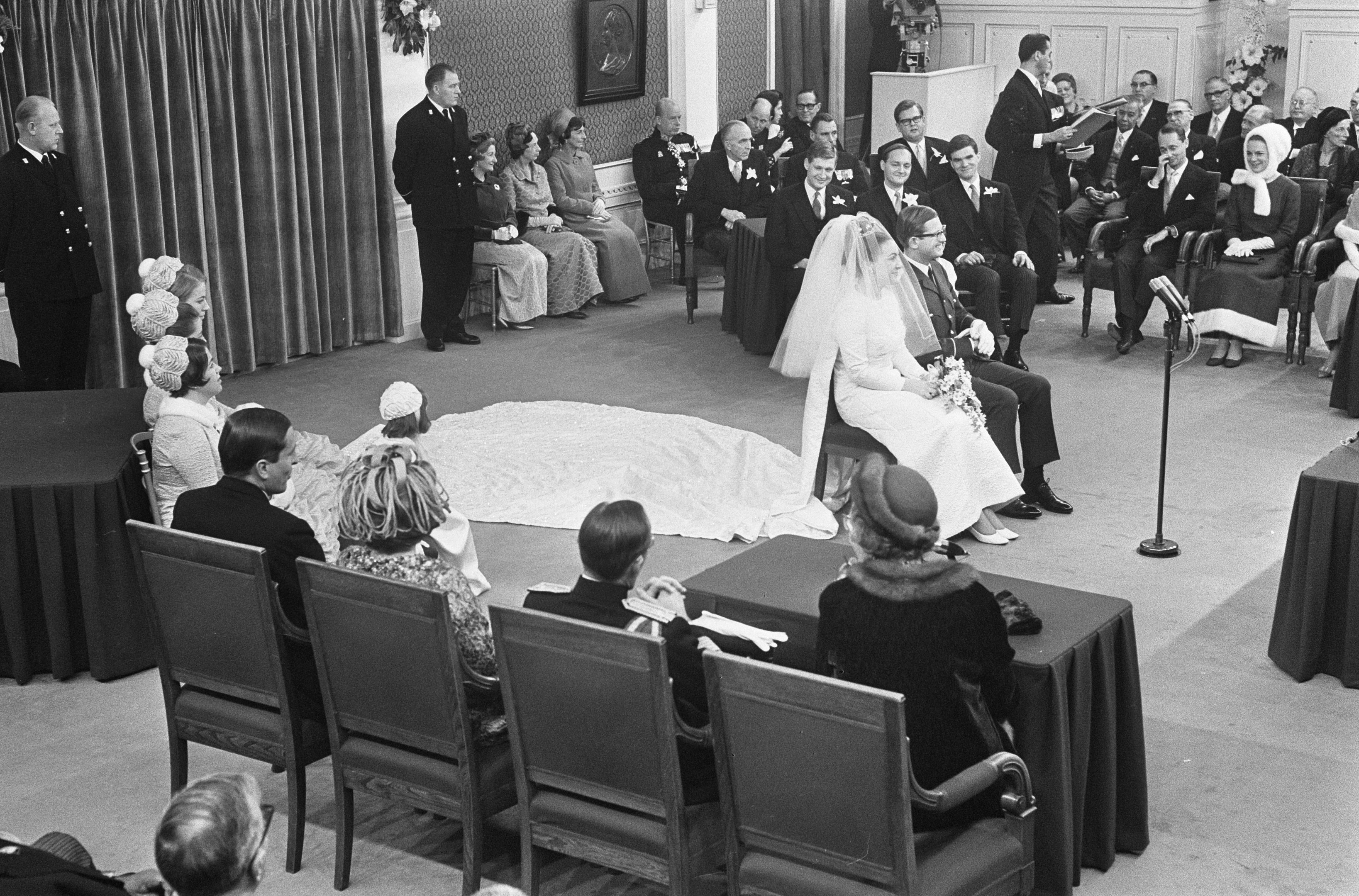 Ein Schwarz-Weiß-Foto einer Hochzeitszeremonie mit Menschen, die sitzen und stehen, einem Mann mit Mikrofon, einer Frau mit Blumenstrauß, einem Podium mit Mikrofon, Blumenschmuck, einem Vorhang, einem gerahmten Foto an der Wand und einer Tür im Hintergrund.