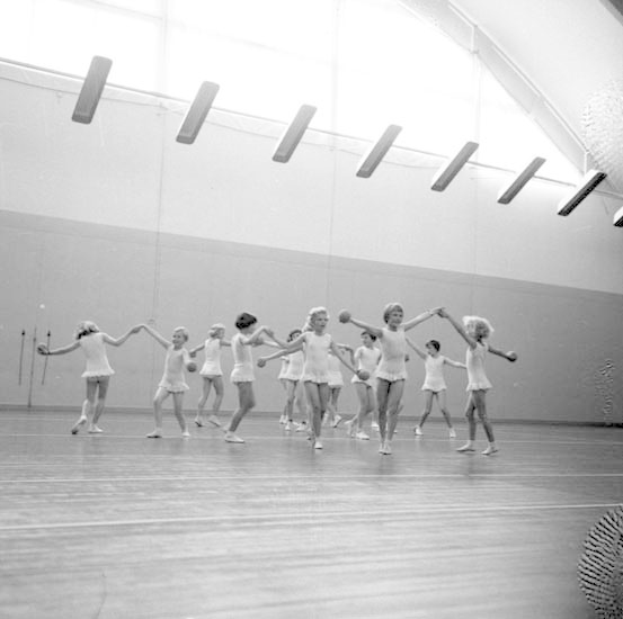Schwarzes und weißes Bild von jungen Mädchen, die sich in einem Studio mit einem Ball auf der rechten Seite, Deckenlichtern und einem Wandobjekt im Hintergrund an den Händen halten und tanzen.