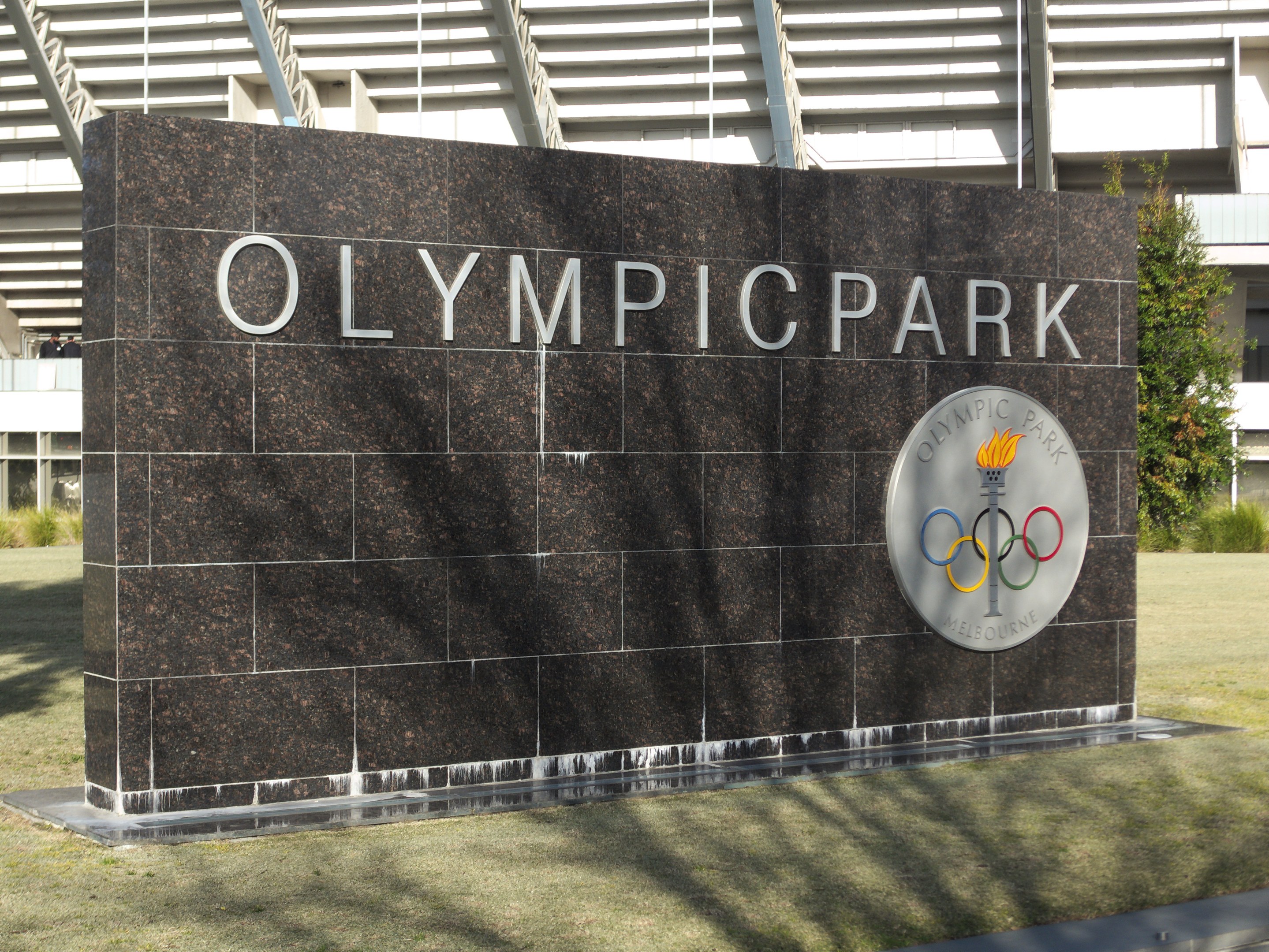 Olympischer Park in Melbourne, Australien, mit einem Namensschild im Vordergrund, Gras, Bäumen und einem entfernten Gebäude.