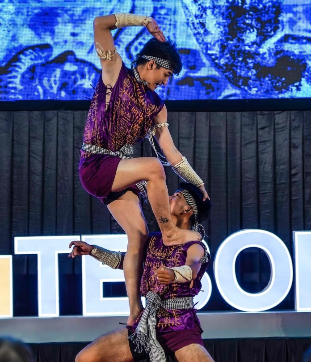 Ein Mann und eine Frau in lila Kleidern führen Akrobatik auf der Bühne vor einem Publikum auf, wobei der Mann die Frau in seinen Armen hält.