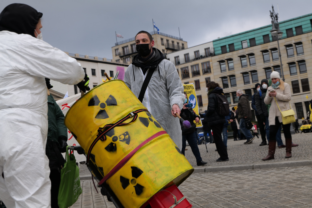 Ein Mann in einem weißen Anzug trägt einen gelben Eimer mit einem radioaktiven Symbol darauf, umgeben von maskierten Individuen mit Taschen, mit Gebäuden, einer Laterne, Fahnen und einem bewölkten Himmel im Hintergrund.