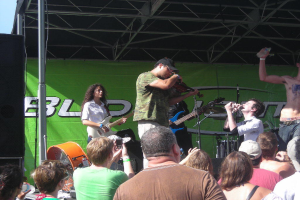 Eine Gruppe von Musikern auf der Bühne, die verschiedene Instrumente spielen, mit einem stehenden Publikum davor, einem großen schwarzen Lautsprecher links und einem grünen Banner im Hintergrund.