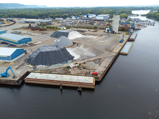 Luftaufnahme eines großen Gewässers mit einem zentralen Dock, umgeben von Gebäuden, Schuppen, Fahrzeugen, Kränen und anderen Strukturen, vor dem Hintergrund von Bäumen, Hügeln und einem klaren blauen Himmel.