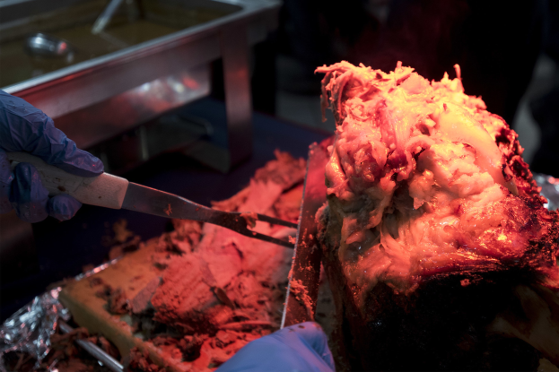 Eine Person mit Handschuhen hält ein Messer über ein Lebensmittel auf einem Tisch, mit zusätzlichen Gegenständen im Hintergrund.