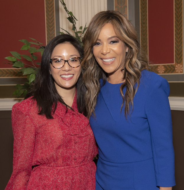 Zwei Frauen, die auf der 2019 Women in Film Pre-Oscars Luncheon zusammen lächeln, eine in einem blauen Kleid und die andere in einem roten Kleid mit Brille, stehen neben einer Pflanze und einem Vorhang.