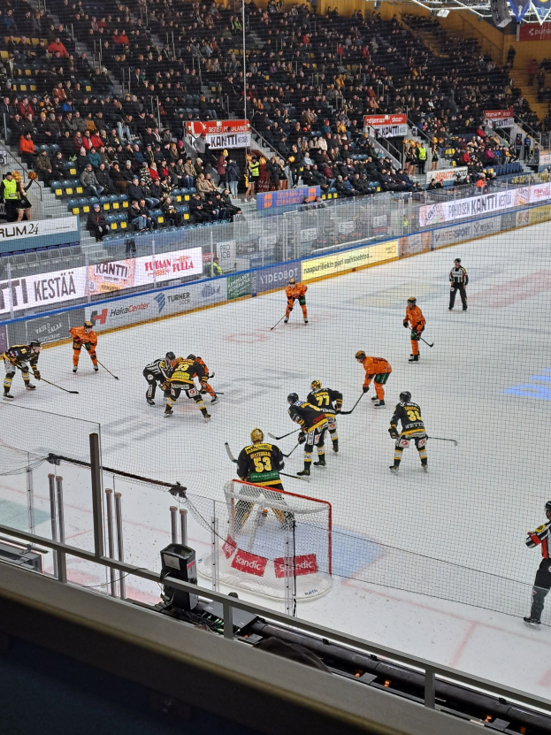 Eine Gruppe von Menschen, die Hockey auf einem Eisring mit einem Netz und einer umgebenden Absperrung spielen, Zuschauern im Hintergrund und Bannern mit Text, unter Stadionbeleuchtung.