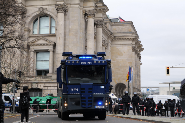 Eine Gruppe von Polizisten steht vor einem großen Gebäude mit Fenstern, Säulen und Bogengängen, während eine Person links eine Kamera hölt und Fahrzeuge auf der Straße vorbeifahren. Trees, traffic signals, flagpoles, and a clear blue sky are visible in the background.