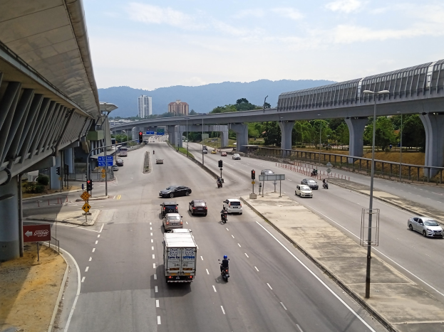 Eine verkehrsreiche Autobahn mit Verkehr, einer Brücke, Straßenlaternen, Verkehrsampeln, Hinweisschildern, Bäumen, Gebäuden, Hügeln und einem bewölkten Himmel im Hintergrund.