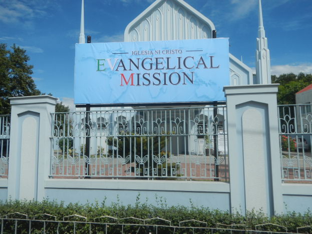 Außenansicht einer Kirche mit einem Metallzaun, einem Schild mit der Aufschrift "Evangelische Mission", umgeben von Bäumen, einem Haus und Pflanzen unter einem bewölkten Himmel.