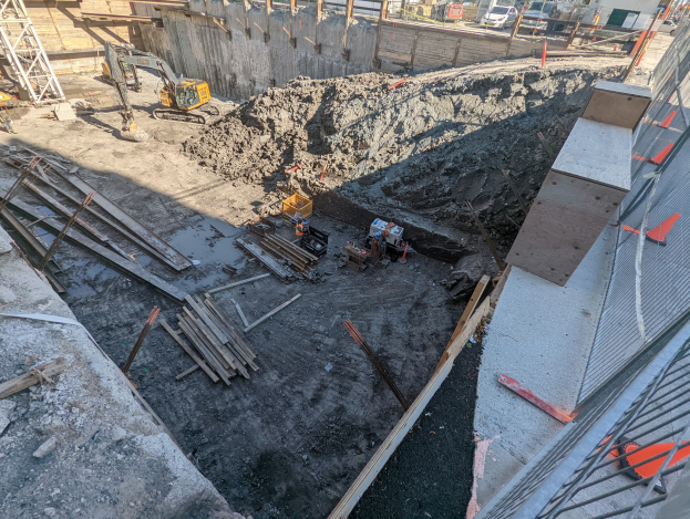 Baustelle mit einem großen Schutthaufen, Kränen, Holzstangen, einem Maschendrahtzaun auf der rechten Seite, Gebäuden, Fahrzeugen, Pfählen, Bäumen und Himmel im Hintergrund.