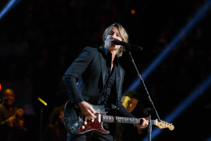 Keith Urban performs on stage playing a guitar with a microphone, accompanied by musicians in a dimly lit setting.