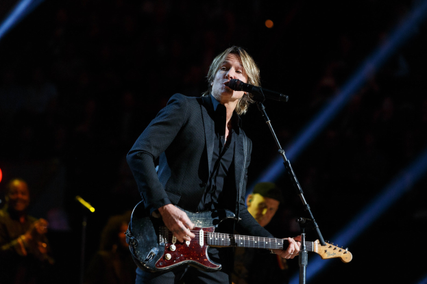 Keith Urban performs on stage playing a guitar with a microphone, accompanied by musicians in a dimly lit setting.
