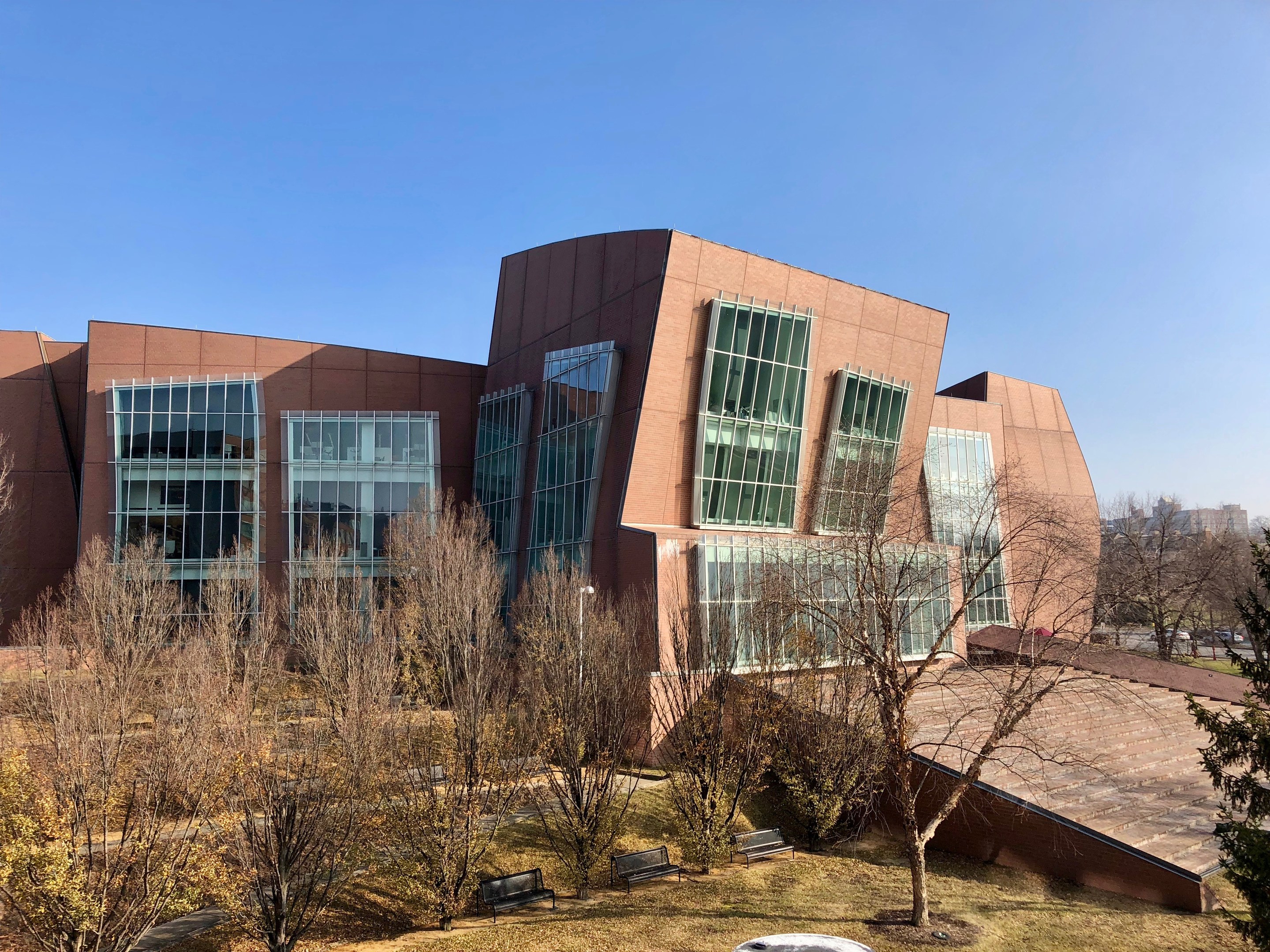Großes mehrstöckiges Gebäude mit zahlreichen Fenstern, umgeben von Bäumen, Bänken, Gras und einem Laternenmast, mit anderen Gebäuden und einem klaren blauen Himmel im Hintergrund.