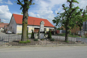 Ein kleines Dorf mit einer Kirche in der Mitte, umgeben von einem Metallzaun, Bäumen, Pflanzen, Gras, einem Schild, Fahrzeugen, Pfosten, Drähten und einem bewölkten Himmel; die Kirche ist die Kirche des Heiligen Grabes in St. Vitus.