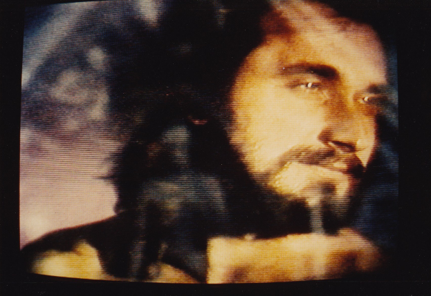 Ein Mann mit langem dunklem Bart und Haar, der ein weißes Hemd trägt, erscheint auf einem Fernsehbildschirm mit dunklem Hintergrund und schaut direkt in die Kamera.