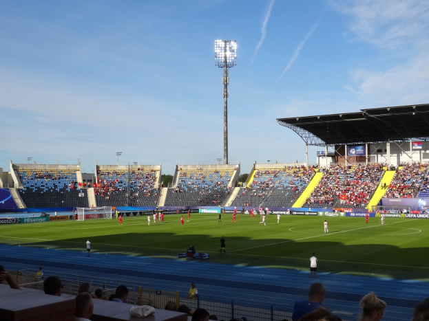 Ein Fötballspiel in einem großen Stadion mit Spielern auf dem Feld und Zuschauern in den Rängen unter Flutlicht, mit dem Himmel im Hintergrund.
