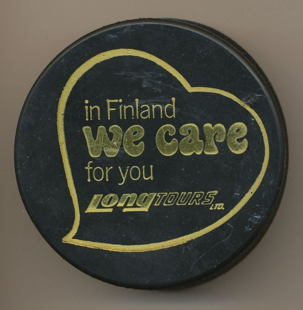 Schwarzer Eishockeypuck mit der Aufschrift "In Finland We Care For You Long Tours" auf einem cremefarbenen Hintergrund.