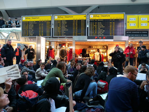 Menschen sitzen und stehen in einem Flughafen während einer Protestaktion, mit Informationsschildern, Puppen in Kleidern und Deckenbeleuchtung im Hintergrund.