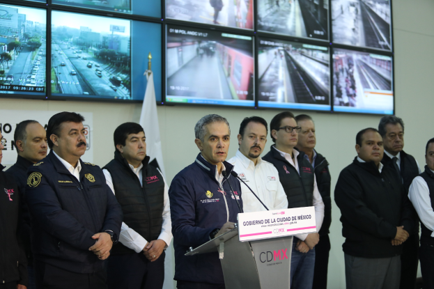 Eine Gruppe von Männern, die vor einem Podium mit einem Mikrofon und einer Tafel mit der Aufschrift "Mexikanische Zoll- und Grenzschutzbehörde" stehen, mit Fernsehgeräten und einer Flagge im Hintergrund.