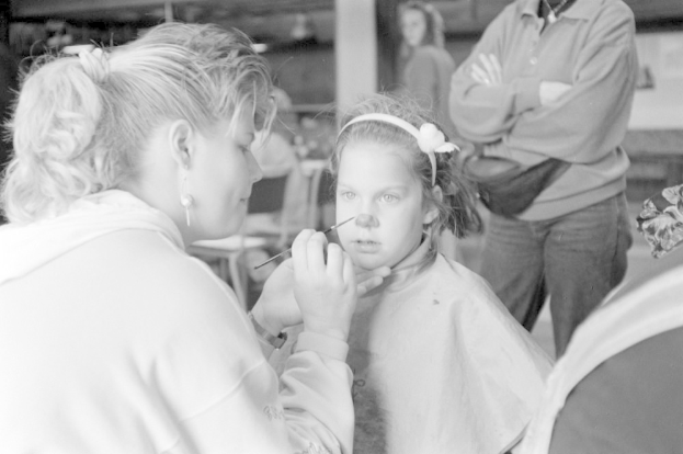 Eine Frau trägt Make-up auf das Gesicht eines jungen Mädchens auf, während zwei andere Personen im Hintergrund stehen.