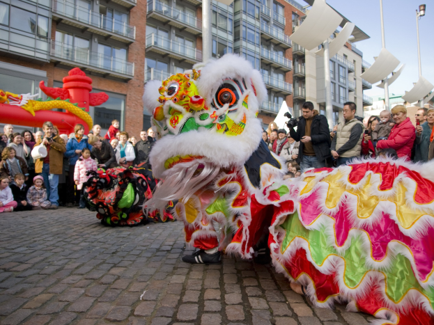Ein lebendiges chinesisches Neujahrsfest in Amsterdam mit einer Löwen-Tanzvorstellung und einer Zuschauermenge, darunter einige mit Kameras, vor einem Hintergrund aus Gebäuden, Laternenmasten und einem klaren blauen Himmel.