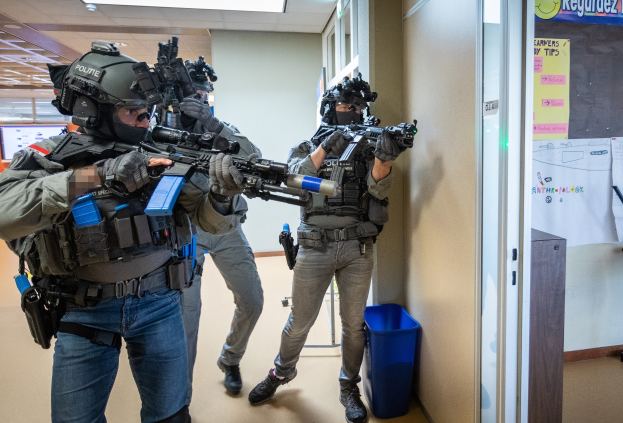 Gruppe von Polizisten in Uniform, die Helme tragen und Waffen halten, steht in einem Raum mit einem Müllcontainer, einer Pinnwand, einer Tür, einem Monitor und Deckenleuchten.
